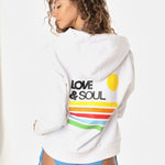 Woman wearing a white hoodie with 'Love & Soul' text and colorful stripes, standing against a white background.